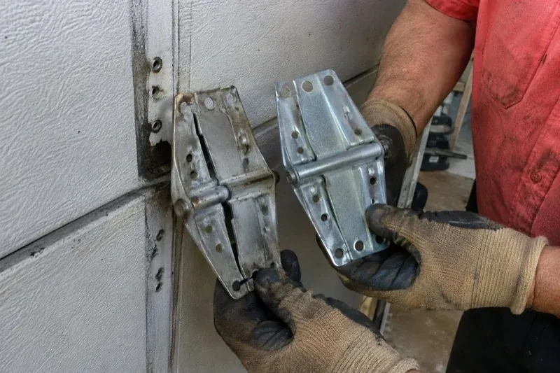 Gloved hands holding two metal garage-door hinges side by side, one discolored with a visible crack and the other bright silver. — Garage Door Hinge Replacement, Whitchurch-Stouffville