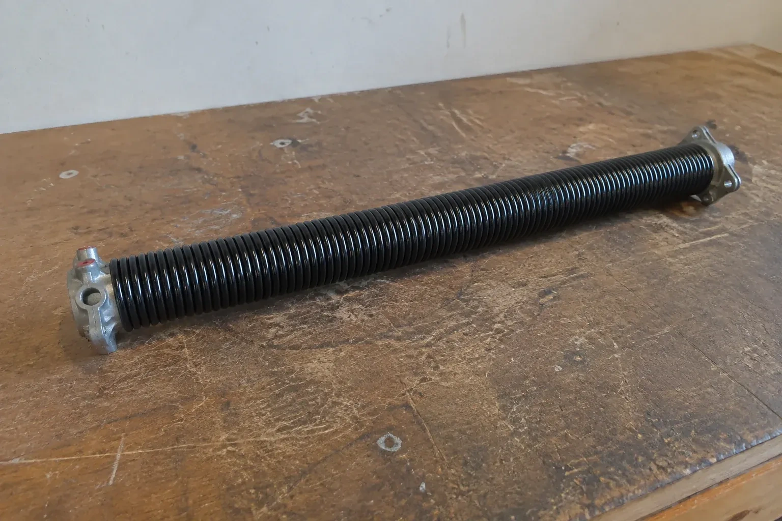 Detached black coil spring with metal end fittings on a workbench. — Torsion Spring Replacement, Whitchurch-Stouffville